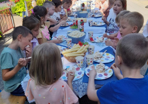 Dzieci siedzą na tarasie przy połączonych stołach i jedzą tort.