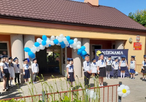 Julka, Antek i Ala stoją na środku tarasu i zapraszają na występ. Po bokach stoją w dwóch półkolach koledzy i koleżanki. W tle budynek przedszkola, dekoracja, balonowa girlanda.