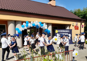 Dzieci tańczą w parach przy piosence. W tle budynek przedszkola oraz dekoracja z okazji pożegnania przedszkola