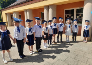 Grupka dzieci stoi w półkolu na tarasie. Dzieci ubrane są na galowo. Na głowach mają granatowe birety, a do bluzek przypięte granatowe kokardy.