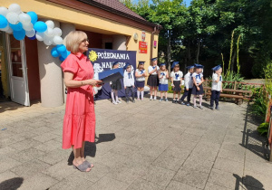 Pani dyrektor stoi na tarasie i składa podziękowania rodzicom. W tle dzieci ubrane na galowo, dekoracja, girlanda balonowa.