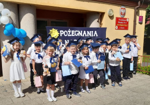 Dzieci z dyplomami i upominkami pozują do wspólnego zdjęcia na tarasie. Przedszkolaki ubrane są na galowo, na głowach mają birety.