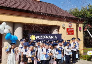 Dzieci z dyplomami i upominkami stoją na tarasie, a nad nimi wiruje confetti.