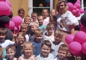 Zdjęcie grupowe dzieci wraz z wychowawcą na schodach przedszkola udekorowanych balonami różowo – czarnymi.
