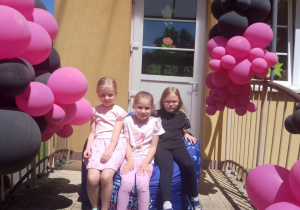 Nikola, Marysia i Zuzia siedzą na pufie na schodach przedszkola udekorowanych balonami różowo – czarnymi.