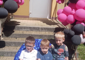 Mikołaj, Adaś i Fabian pozują do zdjęcia siedząc na pufie przed schodami udekorowanymi balonami.