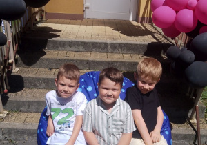 Trzech chłopców pozuje do zdjęcia siedząc na pufie przed schodami udekorowanymi balonami.