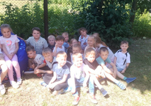 Zdjęcie grupowe dzieci w ogrodzie przedszkolnym. Przedszkolaki siedzą na macie i pufie.