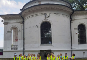 Dzieci ubrane w kamizelki odblaskowe stoją obok siebie. W tle Muzeum Bitwy nad Bzurą oraz armata.