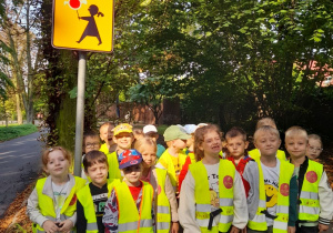 Dzieci ubrane w kamizelki odblaskowe stoją przed znakiem drogowym "Agatka'".