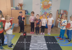 Zabawa tematyczna "Ruch drogowy". Na dywanie leży mata przedstawiająca ulicę, a na niej stoi model sygnalizacji świetlnej. Obok stoją dzieci w roli kierowców, pieszych i policjanta.