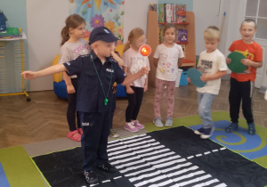 Chłopiec w stroju policjanta stoi na macie przedstawiającej ulicę i kieruje ruchem drogowym. Obok dzieci w roli kierowców i przechodniów.