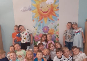 Dzieci pozują do wspólnego zdjęcia. W tle dekoracja przedstawiająca słonko, kwiatki, wiszące chmurki oraz napis "Słoneczka".