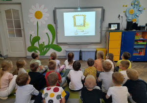 Dzieci siedzą na dywanie przed tablicą multimedialną i oglądają bajkę pt. "Kropka".