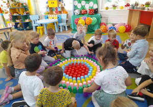 Dzieci siedzą na dywanie i układają z kolorowych kubków kropkę.