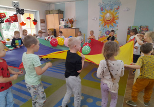 Dzieci poruszają się w kole przy muzyce trzymając chustę animacyjną, na której leżą balony w kropki.