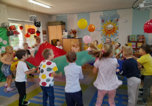 Dzieci stoją przy chuście animacyjnej i podrzucają do góry balony.