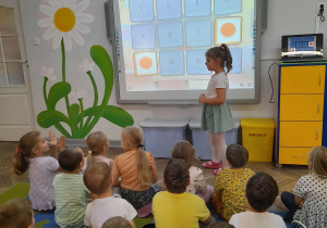 Dzieci siedzą na dywanie, a przed tablicą multimedialną stoi dziewczynka, która podczas zabawy "Memory - kolorowe kropki" odkryła dwie takie same kropki.