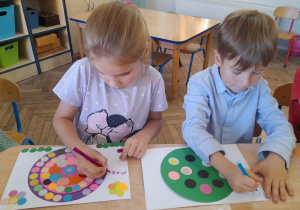 Dwoje dzieci siedzi obok siebie przy stole i próbuje pisać swoje imię na pracy plastycznej.