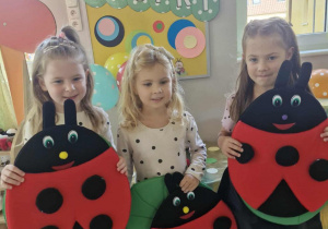 Ola, Alicja i Oliwia trzymają w dłoniach duże biedronki z przyklejonymi kropkami. W tle widać dekorację przygotowaną z okazji Międzynarodowego Dnia Kropki.