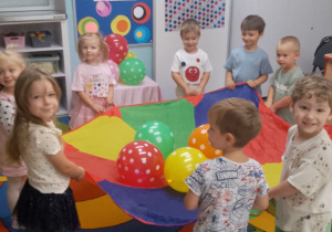 Dzieci poruszają się w kole przy muzyce trzymając chustę animacyjną, na której leżą balony w kropki.