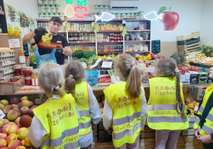 Cztery dziewczynki stoją przed ladą w warzywniaku, a Pan właściciel wkłada do torebki winogron.