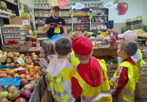 Dzieci stoją obok lady i oglądają owoce, warzywa oraz inne produkty na półkach. Pan właściciel warzywniaka wkłada do torby banany.