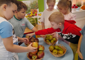 Grupka dzieci w stoi w sali obok stolika i układa w koszyczkach jabłka i gruszki.