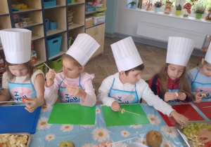 Grupka dzieci siedzi przy stole i w skupieniu nadziewa owoce na patyczki.