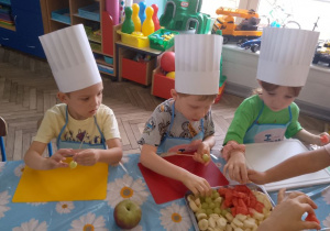 Grupka dzieci siedzi przy stole i wykonuje owocowe szaszłyki.