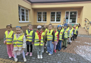 Dzieci ubrane w kamizelki odblaskowe stoją przed wejściem do przedszkola.
