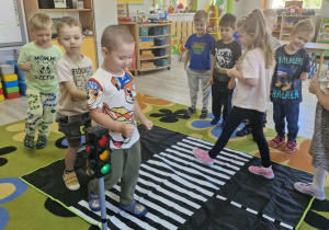Dzieci w trakcie zabawy dotyczącej bezpieczeństwa na drodze. Na dywanie leży mata przedstawiająca ulicę, a na niej stoi model sygnalizacji świetlnej. Obok stoją dzieci wcielające się w role kierowców oraz pieszych.