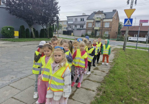 Dzieci ubrane w kamizelki odblaskowe idą w parach po chodniku.