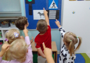 Dzieci stoją przed tablicą, na której są ilustracje świateł drogowych i przejścia dla pieszych.
