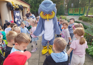 "Słoneczka" ustawione w kole, tańczą z maskotką sowy. W oddali widać balony w kolorze złotym i czarnym.