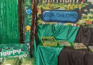 Dekoracja z okazji Dnia Chłopaka: na tablicy znajduje się Minecraftowy baner z napisem Dzień Chłopca, na szafce pokrytej zielonym i brązowym materiałem leżą zielone koszyczki z upominkami dla dzieci. Nad tablicą wiszą kwadratowe balony w kolorze zielonym i brązowym.