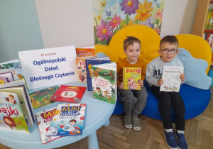 Mikołaj i Bartek siedzą na kanapie obok stolika z książkami i napisem "Ogólnopolski Dzień Głośnego Czytania". Chłopcy trzymają książki zaprezentowane przez rodzica.