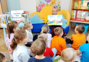 Dzieci siedzą na dywanie, a przed nimi na kanapie Tosia. Dziewczynka prezentuje obrazki w książce. Obok znajduje się Kącik Książki i stolik z książkami.