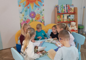 Dzieci siedzą przy stoliku i oglądają książki z przedszkolnej biblioteki. W tle Kącik Książki.