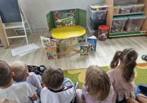 Dzieci siedzą w półkolu na dywanie, przed nimi na stoliku widać rozłożony teatrzyk.