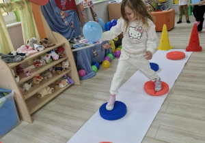 Na podłodze leży biały pas papieru symbolizujący rzekę i kolorowe krążki symbolizujące kamienie. Weronika przeprawia się przez rzekę idąc po kamieniach.