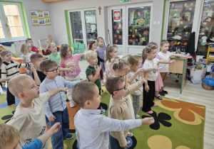 Przedszkolaki stoją na dywanie z wyciągniętą do przodu ręką w trakcie tańca z pokazywaniem.