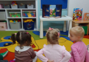 "Pszczółki" siedzą na dywanie przed Teatrzykiem Kamishibai z bajką "Trzy małe świnki". Z tyłu tablica z napisem "Dzień Głośnego Czytania".