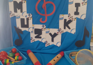 Dekoracja z okazji Dnia Muzyki: na niebieskim materiale przypięty napis Dzień Muzyki i kolorowe nuty. Na szafce leżą instrumenty muzyczne.