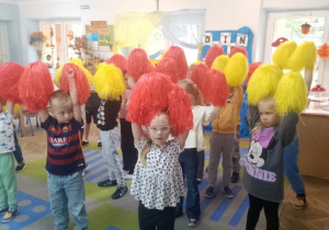 Dzieci stoją w rozsypce i trzymają żółte i czerwone pompony nad głowami.