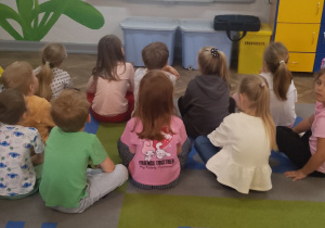 Dzieci siedzą na dywanie przed tablicą multimedialną i oglądają bajkę edukacyjną o instrumentach muzycznych.