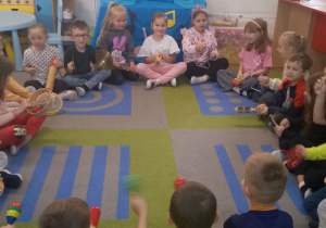 Przedszkolaki siedzą w kole na dywanie i trzymają różne instrumenty muzyczne. W tle dekoracja z okazji Dnia Muzyki.