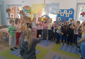 Dzieci stoją w półkolu i naśladują ruchem instrumenty muzyczne, przed nimi stoi dziewczynka i dyryguje trzymając w ręku pałeczkę.