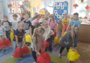Przedszkolaki stoją w rozsypce i trzymają żółte i czerwone pompony.