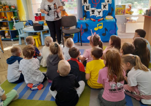 Dzieci siedzą na dywanie, a przed nimi stoi zaproszony gość – mama Bartka. W tle dekoracja z okazji Dzień Muzyki: na niebieskim materiale przypięty jest napis Dzień Muzyki i kolorowe nutki. Na szafce leżą instrumenty perkusyjne.